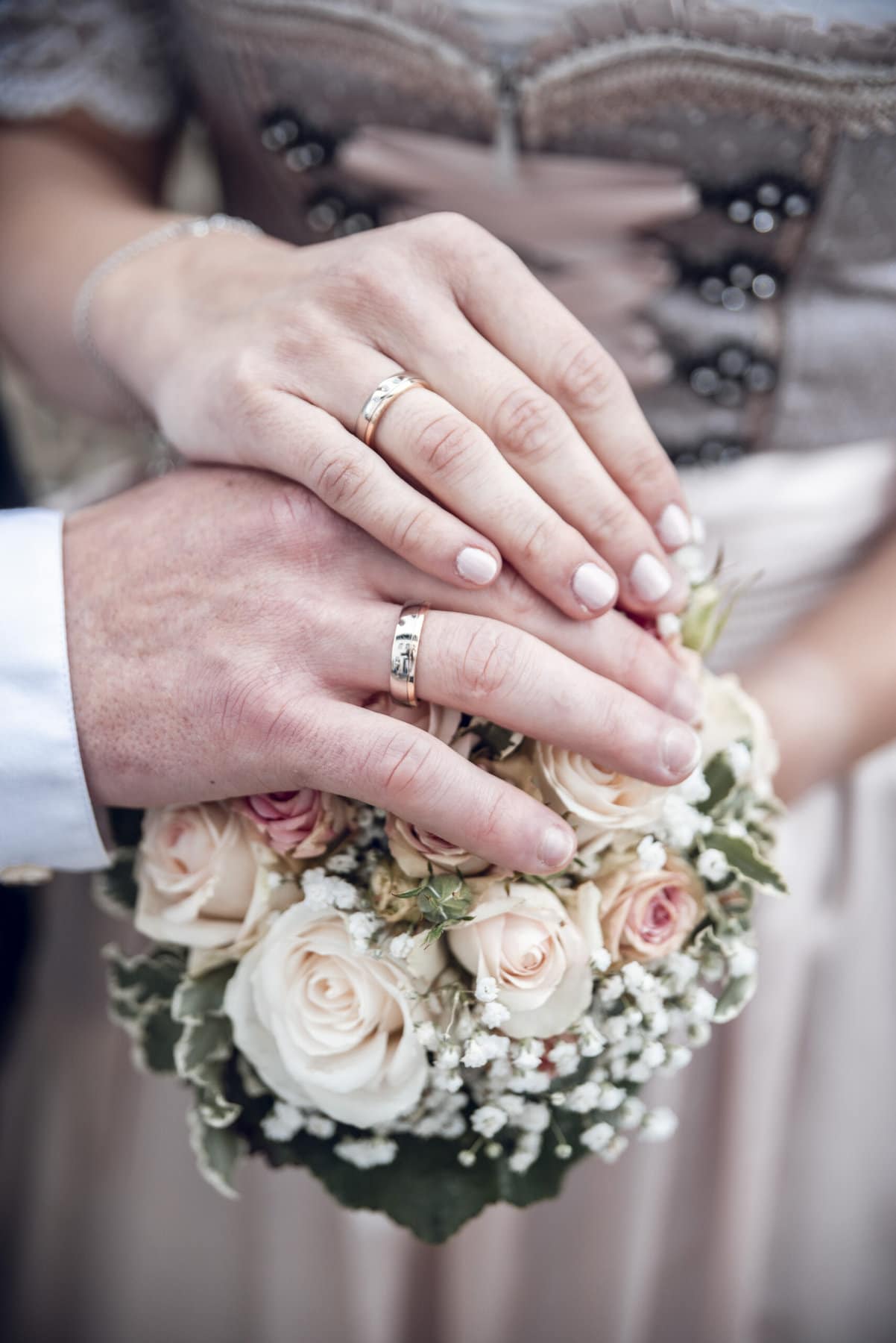 Die Hände des Brautpaars mit Ringen auf dem Braustrauß vor dem Dirndl der Braut.
