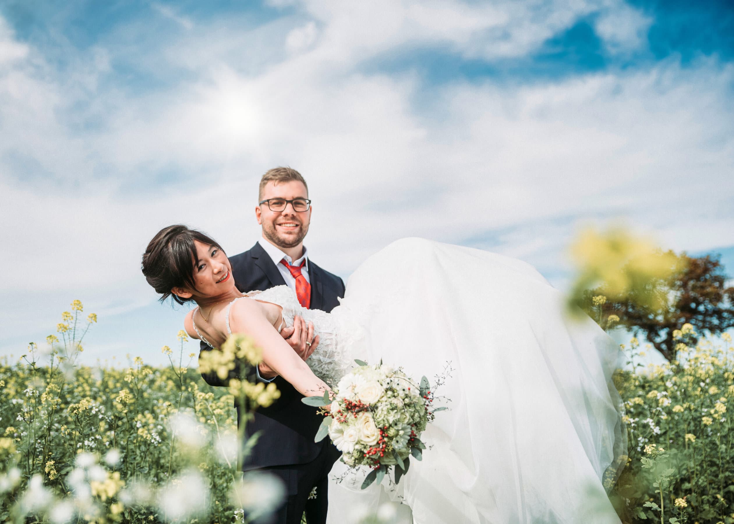 Ein Hochzeitsfoto im Rapsfeld bei Sonnenschein. Der Bräutigam trägt die Braut auf den Händen.