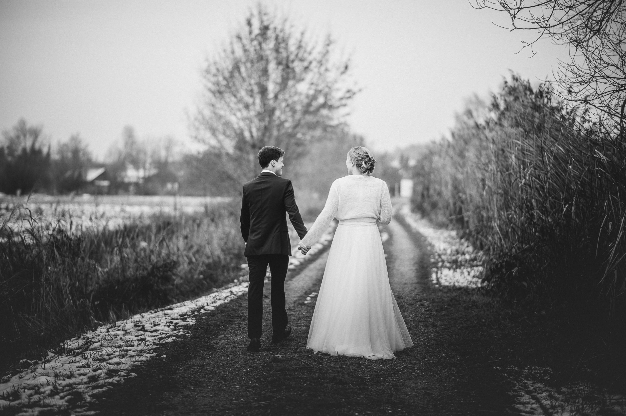 Eine Aufnahme in schwarz-weiß und von hinten. Das Brautpaar läuft Hand in Hand am Klosterweiher in Thierhaupten.