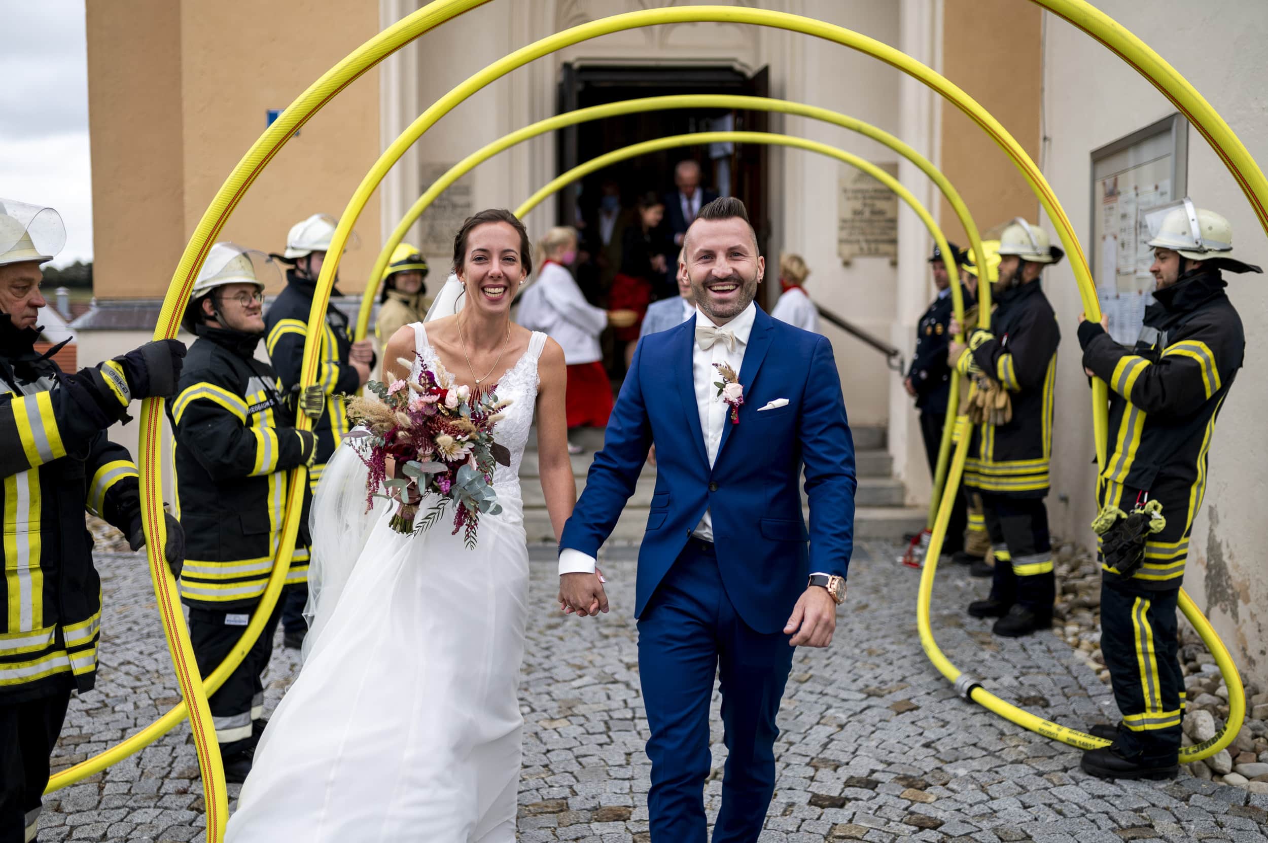 Die Feuerwehr steht bei Hochzeit vor Aindlinger Kirche Spalier und das Brautpaar läuft durch die Bögen aus Feuerwehrschläuchen.