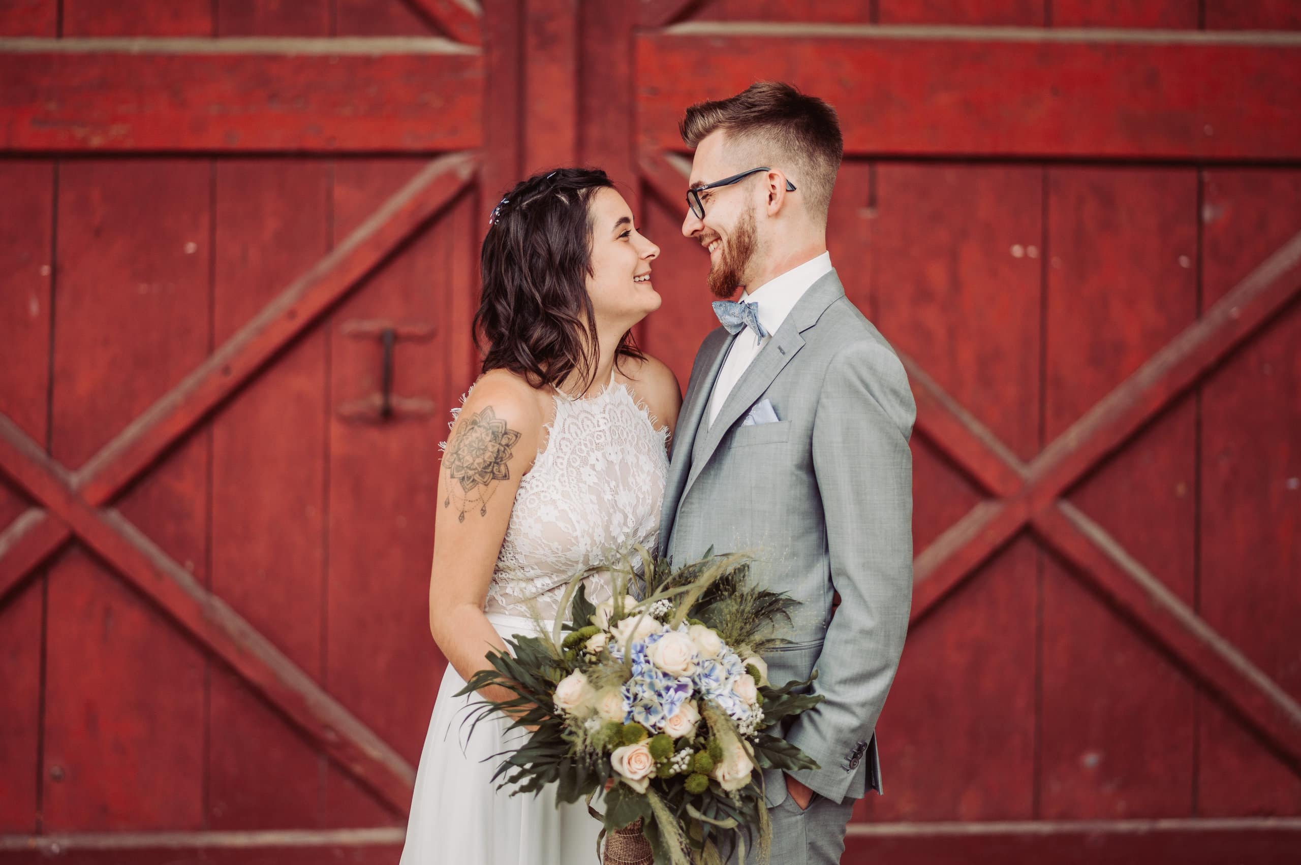 Braut und Bräutigam lächeln einander ins Gesicht vor einem großen roten Holztor.