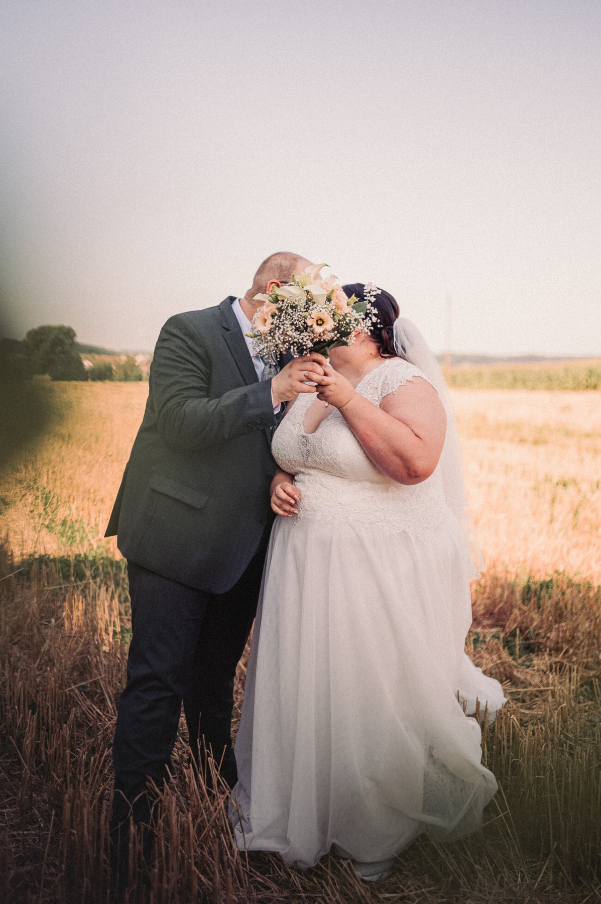 Das Brautpaar küsst sich beim Hochzeitsshooting in einem Feld in Gablingen und verdecken ihr Gesicht mit dem Brautstrauß.