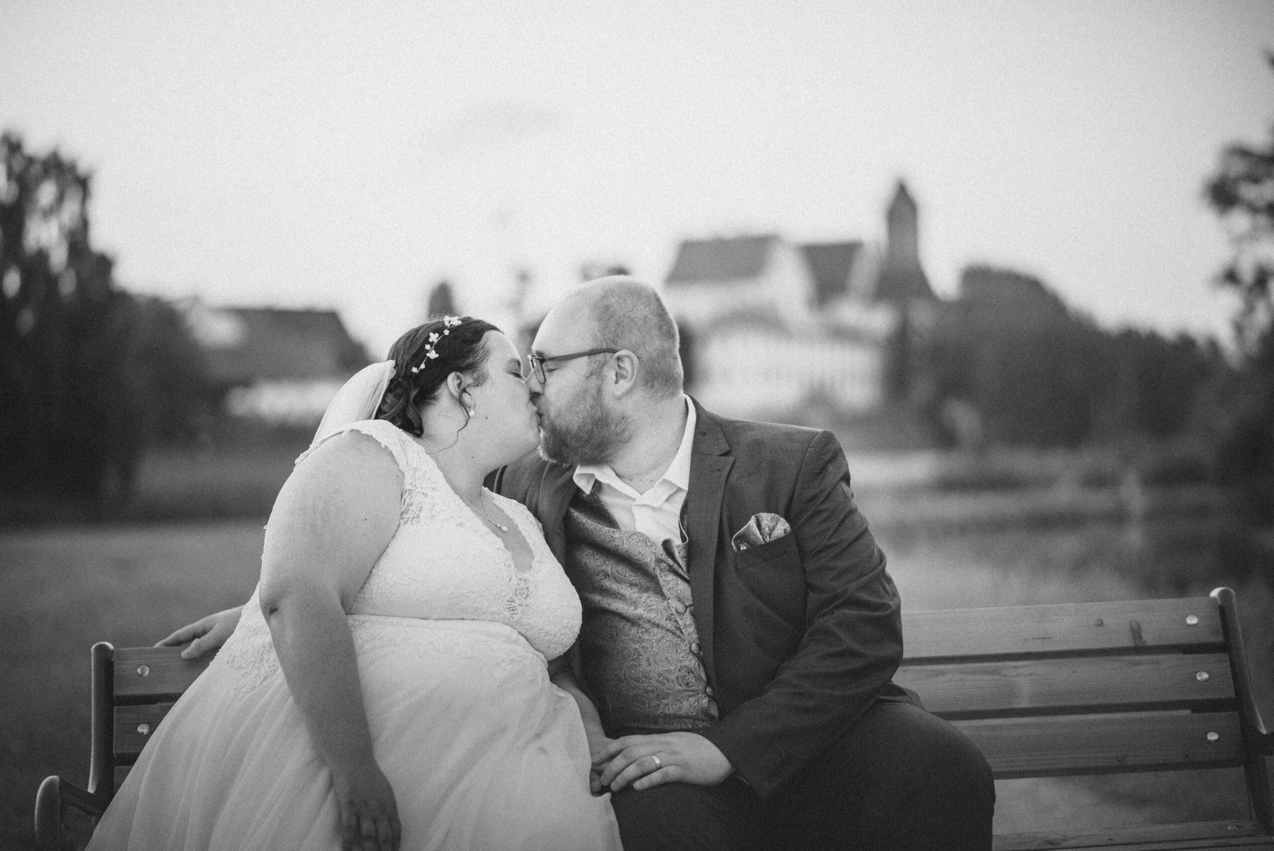 Das Brautpaarshooting im Sonnenuntergang in schwarz-weiß in Gablingen. Das Brautpaar küsst sich auf einer Sitzbank.