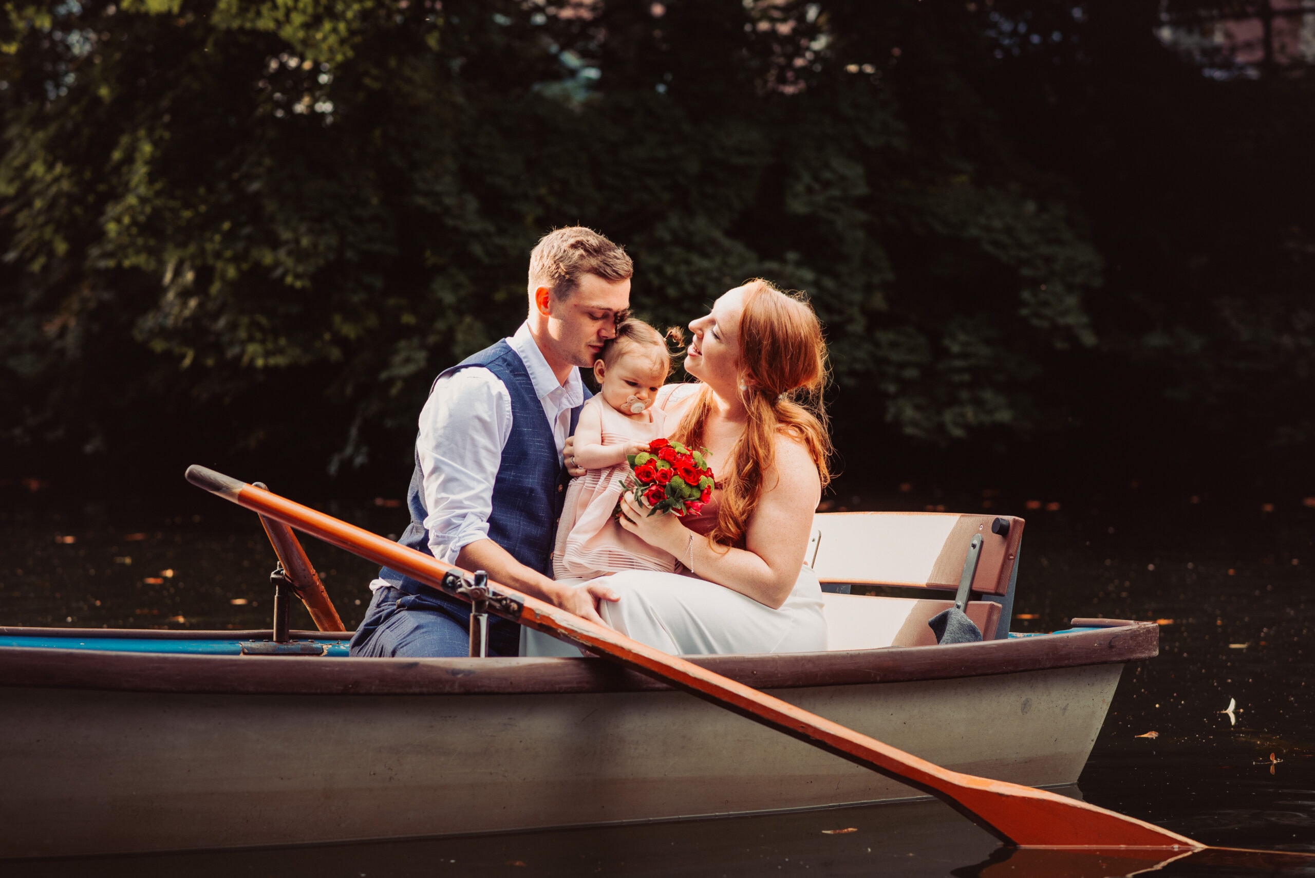 Das After Wedding Shooting in Augsburg. Das Brautpaar sitzt mit ihrem Kind in einem Ruderboot.