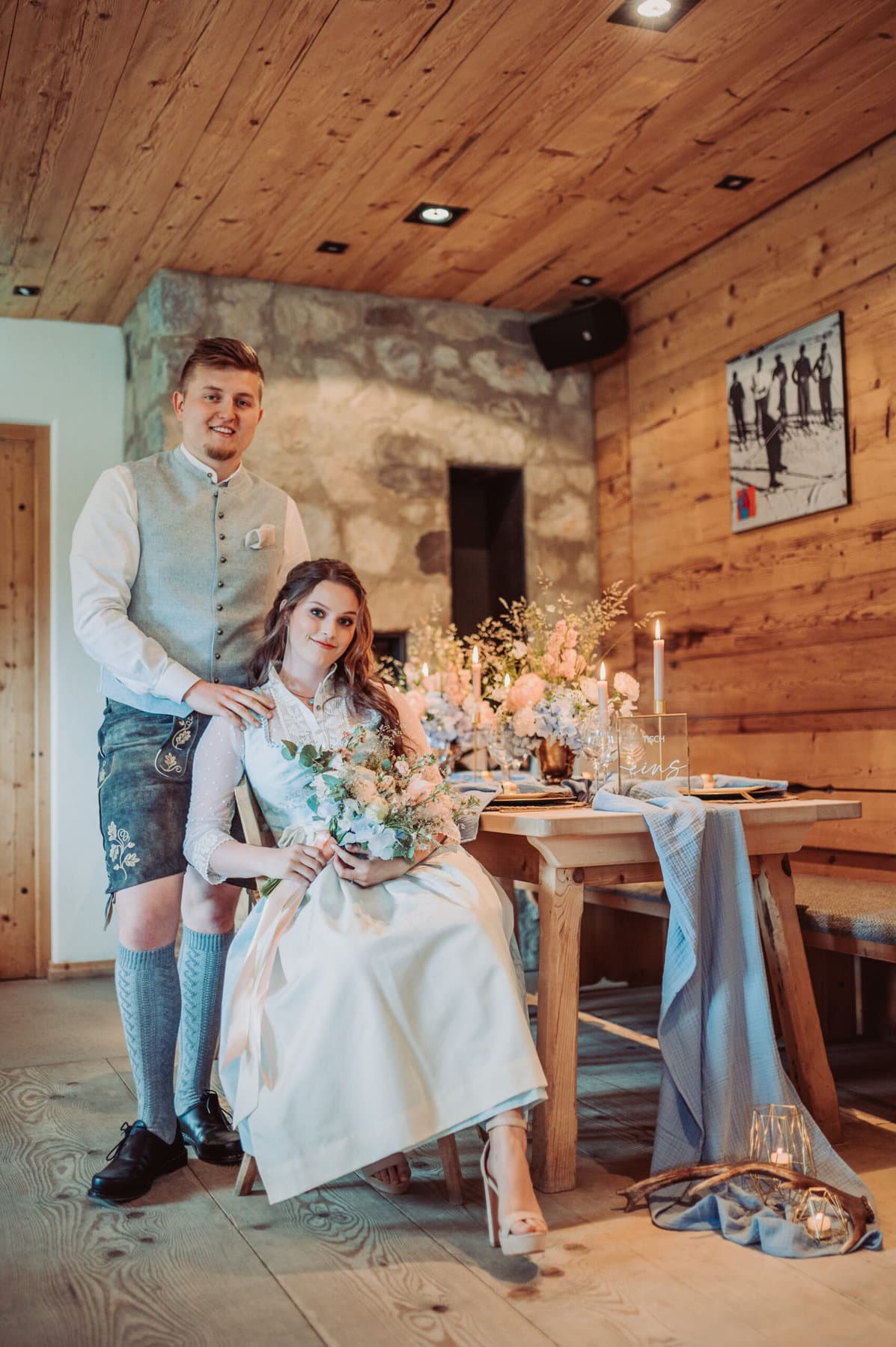 Der Esstisch der Hochzeit auf der Meckatzer Sportalp. Die Braut sitzt an einem gedeckten Tisch und hinter ihr steht der Bräutigam, dabei liegen seine Hände auf ihren Schultern.