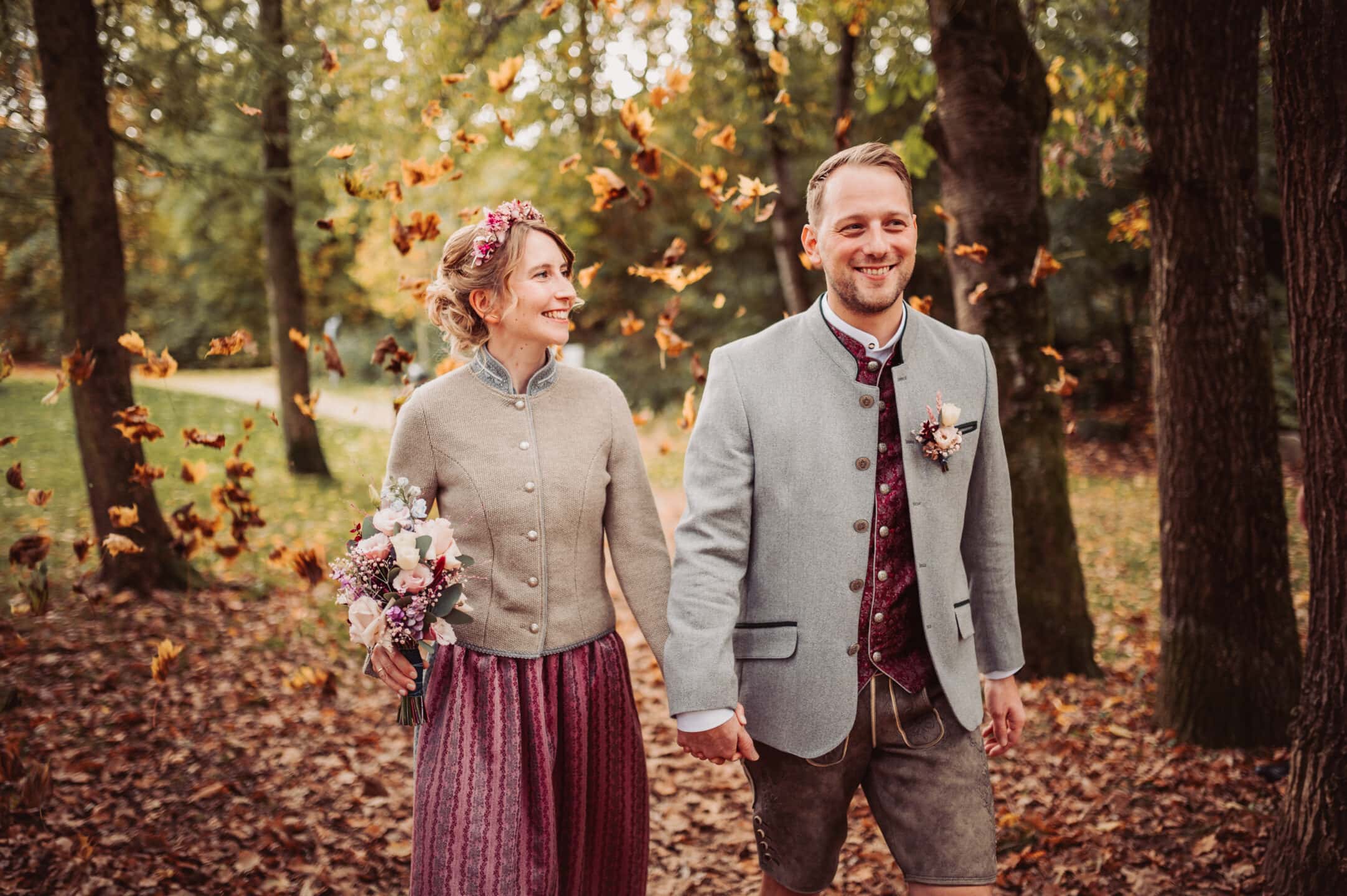 Das Brautpaar läuft beim herbstlichen Brautpaarshooting im Schmutterpark in Neusäß spazieren und die Blätter fallen vom Baum.