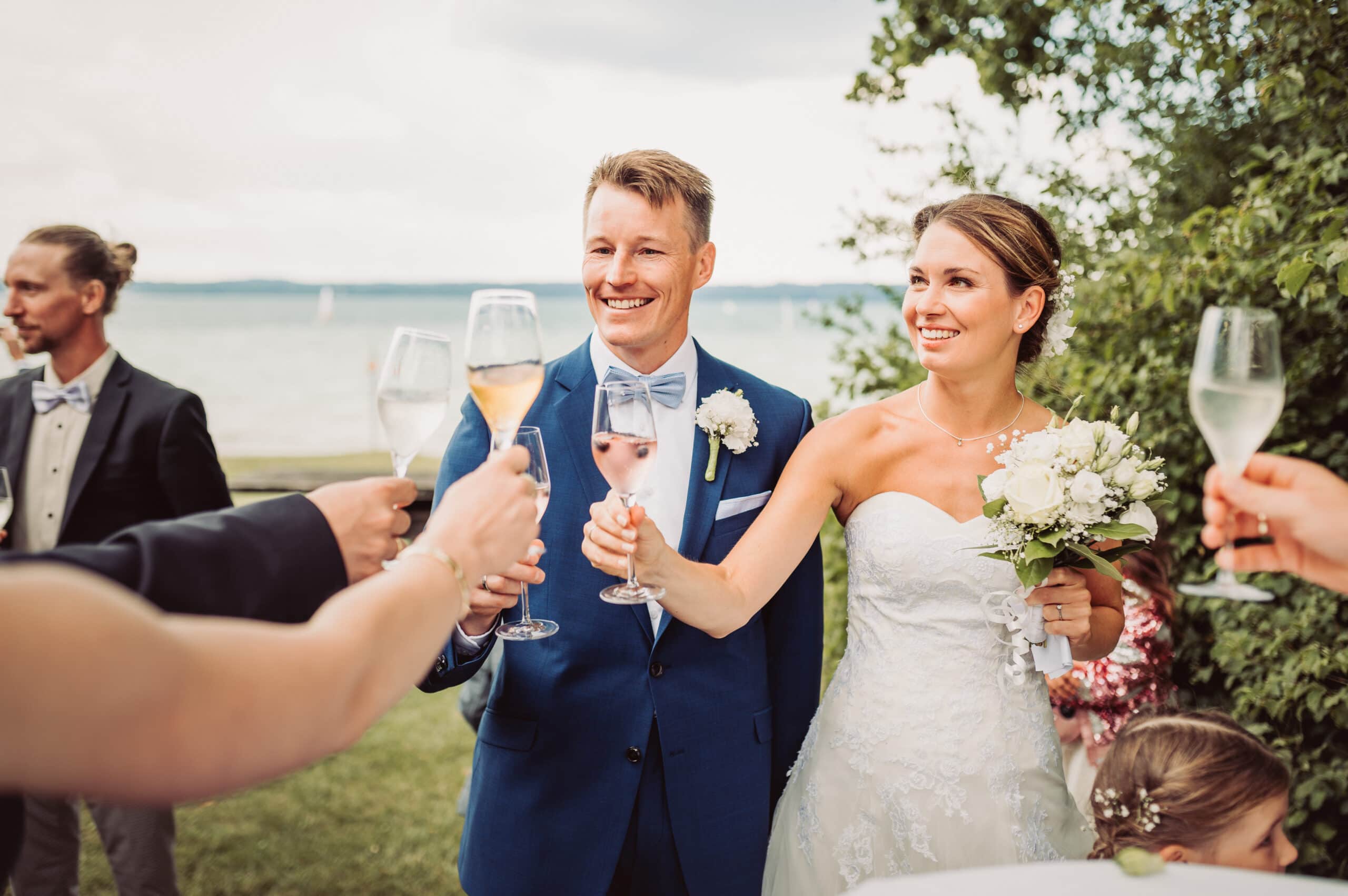 Das Brautpaar stößt bei der Hochzeitsfeier am Starnberger See mit Sekt mit ihren Gästen im Garten vom Seewirt Buchscharner an.