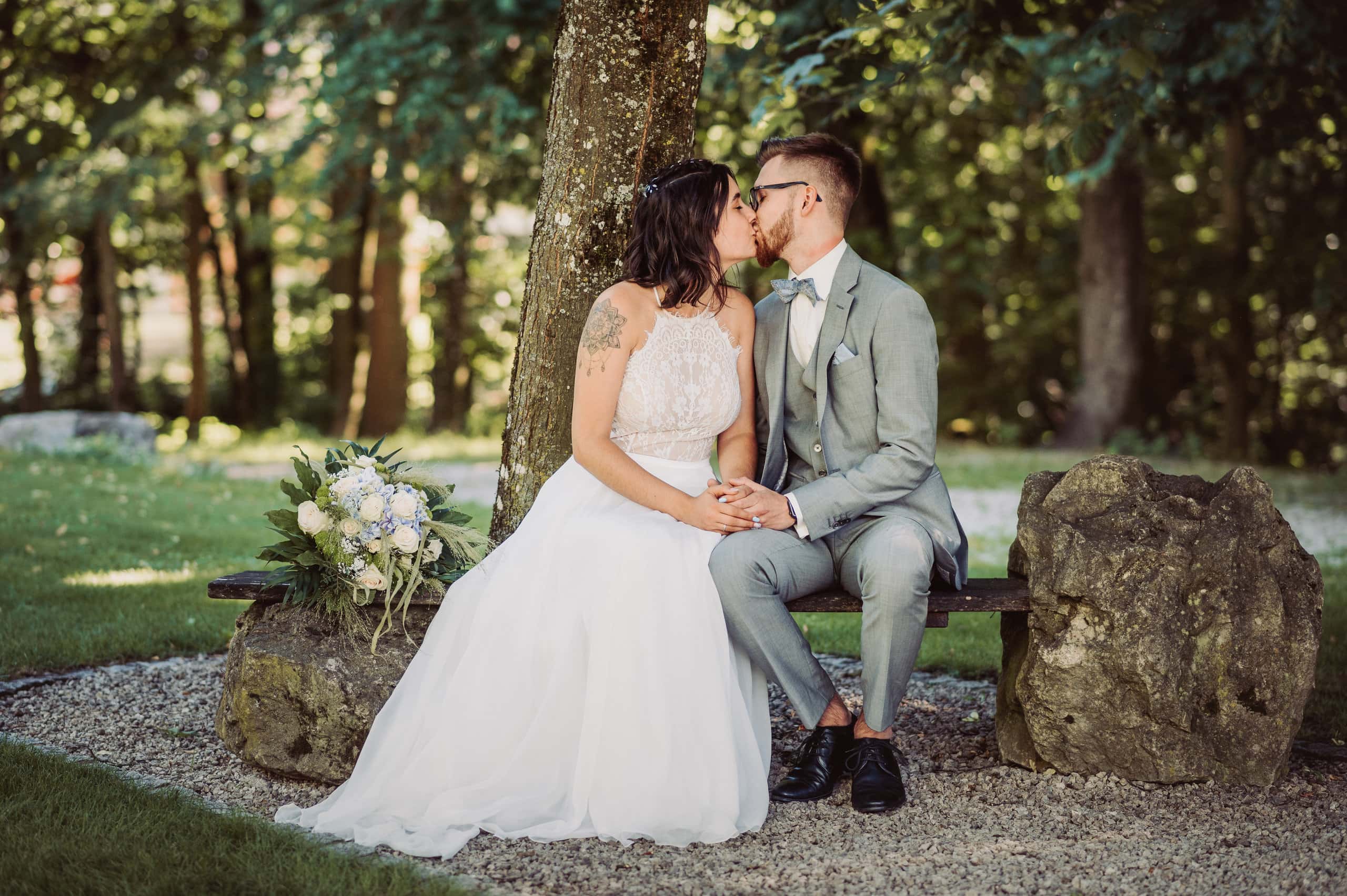 Brautpaar küsst sich auf einer Bank am Waldrand